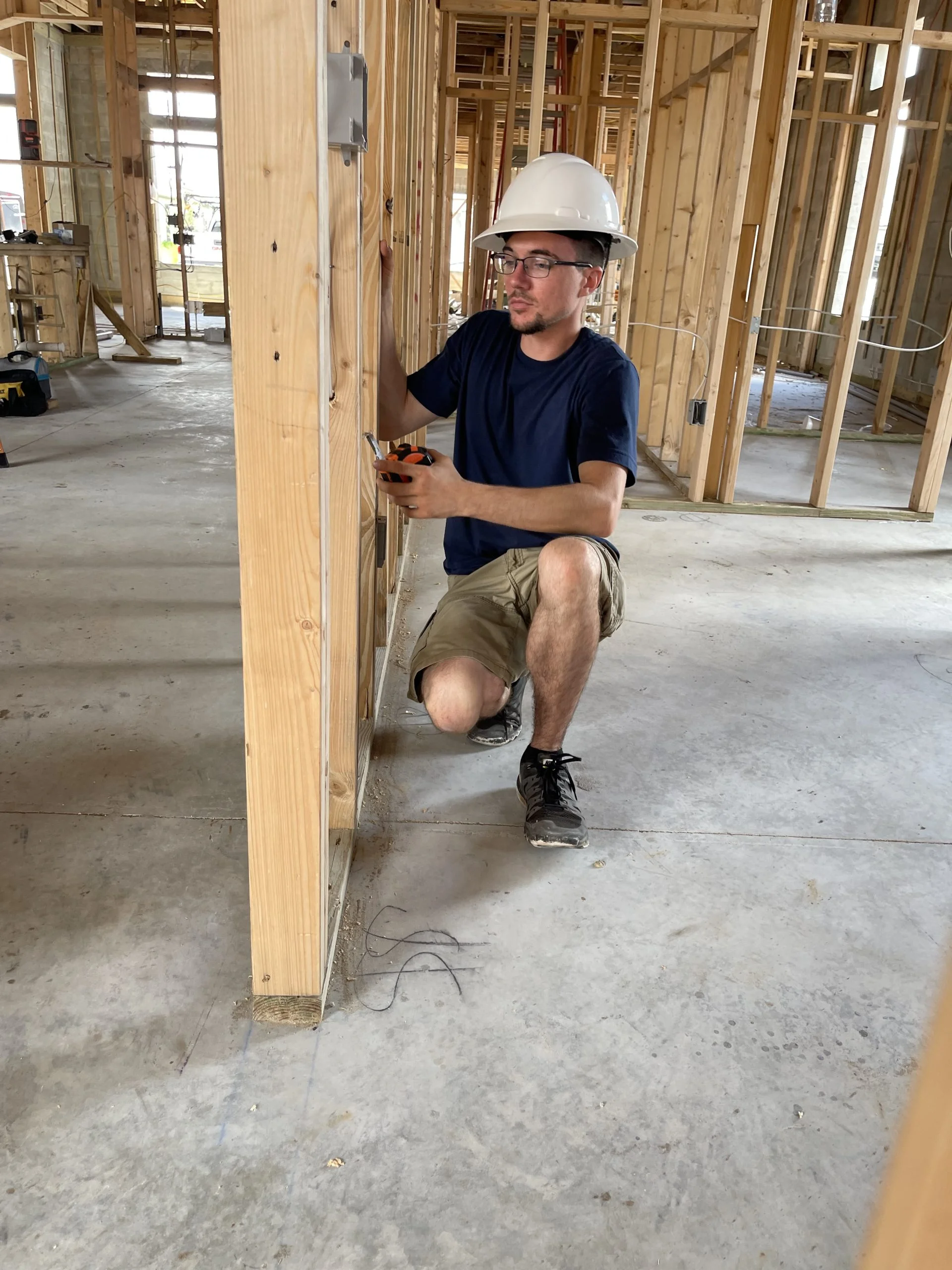 Electrician working on wiring installation in a construction site, wearing a hard hat and measuring tools, surrounded by wooden framing and electrical components, reflecting New Dawn Electric's commitment to skilled craftsmanship.