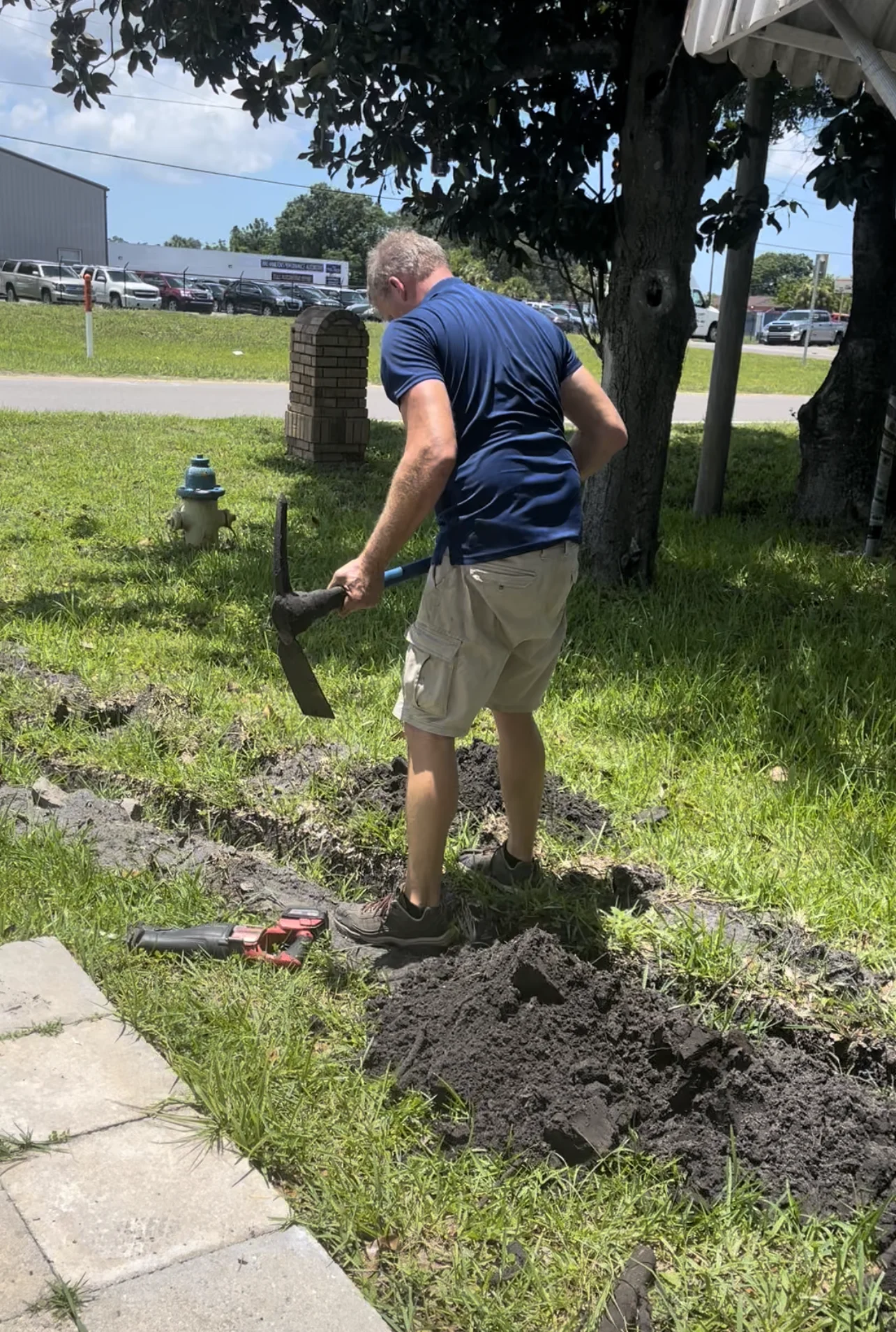 Man digging in grassy area with pickaxe, surrounded by electrical tools, nearby fire hydrant, showcasing hands-on work in electrical service installation.