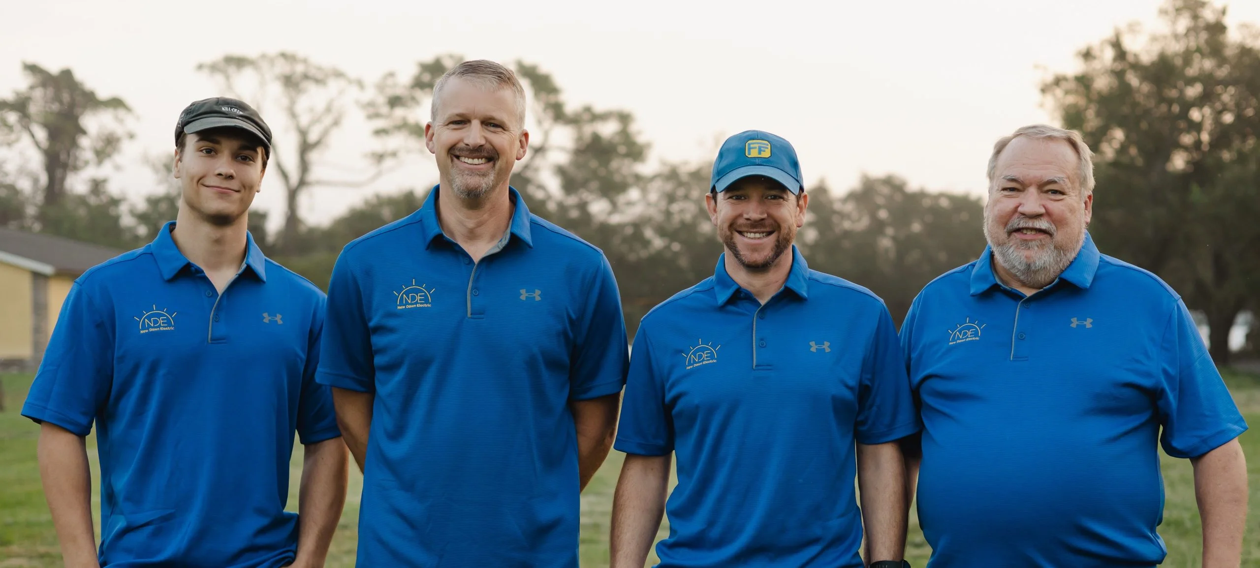 Four male team members of New Dawn Electric smiling in blue polo shirts, showcasing camaraderie and professionalism, set against a natural outdoor background.