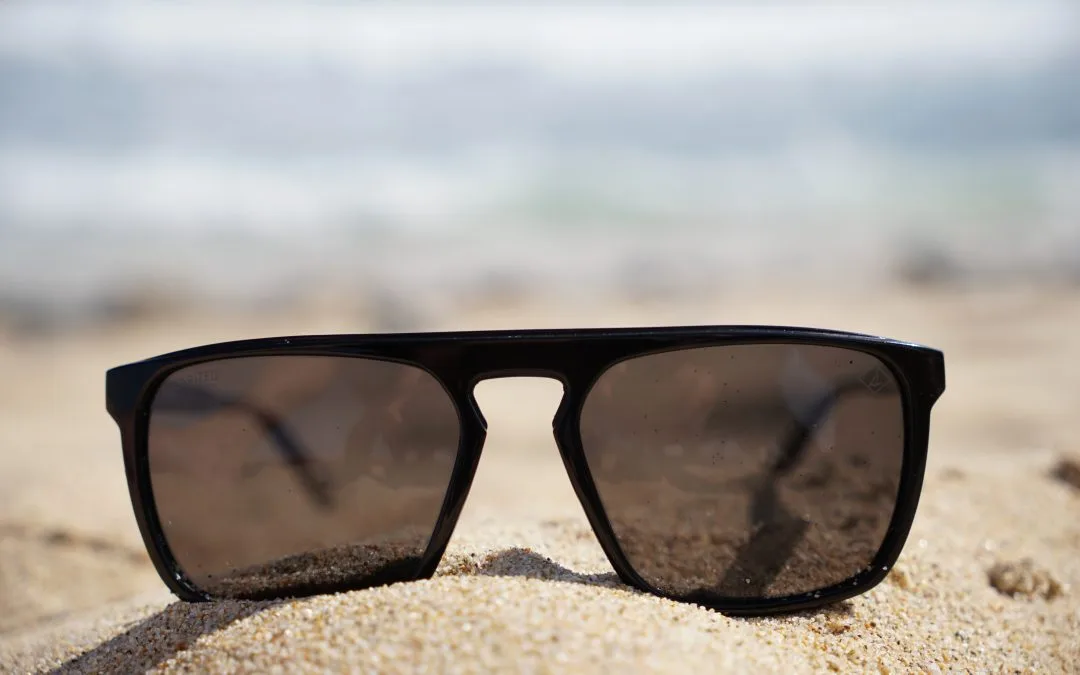Black sunglasses resting on sandy beach, with blurred ocean in background, symbolizing summer comfort and outdoor living in Florida.